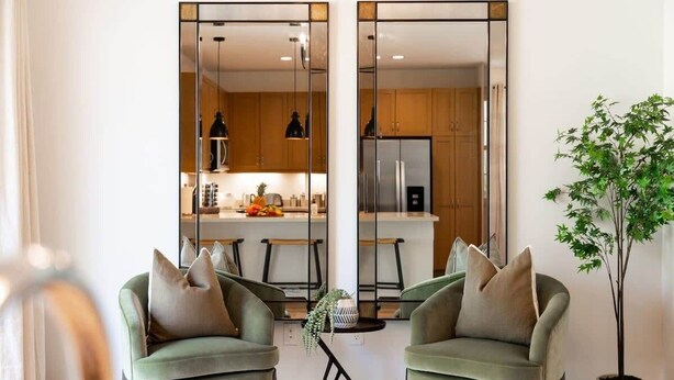 A modern living room with two green armchairs, a small coffee table, and a potted plant. A large mirror reflects a kitchen with wooden cabinets and a refrigerator.