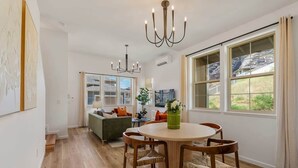 A dining area with a round table and chairs, a chandelier, and large windows with a view of greenery.