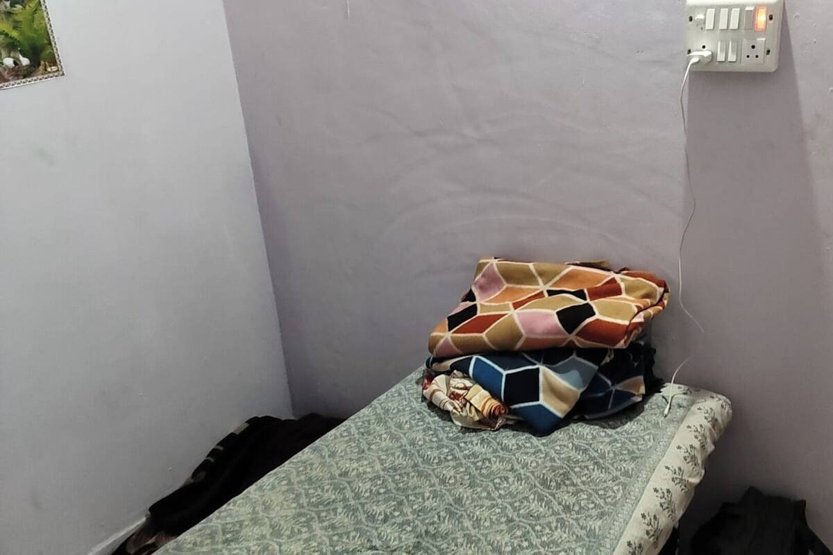Una habitación pequeña y estrecha con una cama individual, un enchufe en la pared y una puerta que se ve a medias.
