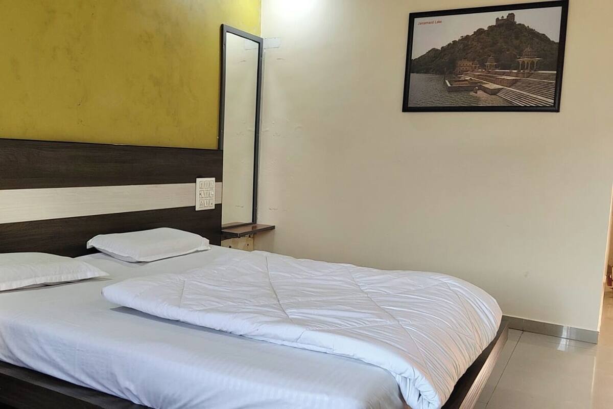 Una cama bien hecha con sábanas blancas en una habitación con pared amarilla y un cuadro de un lago.
