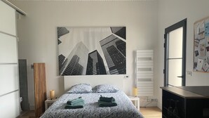 A bedroom with a bed, a large cityscape painting on the wall, a wooden slat, a bedside table with a lamp, and a dresser with wine glasses. - On the road to the castles (Soussans)
