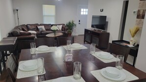 A modern living room with a dining area, featuring a brown leather sofa, a TV, and a dining table set with white plates and glasses. - Newly renovated home near TMC, Downtown, UH, TSU available for short term rental (Houston)