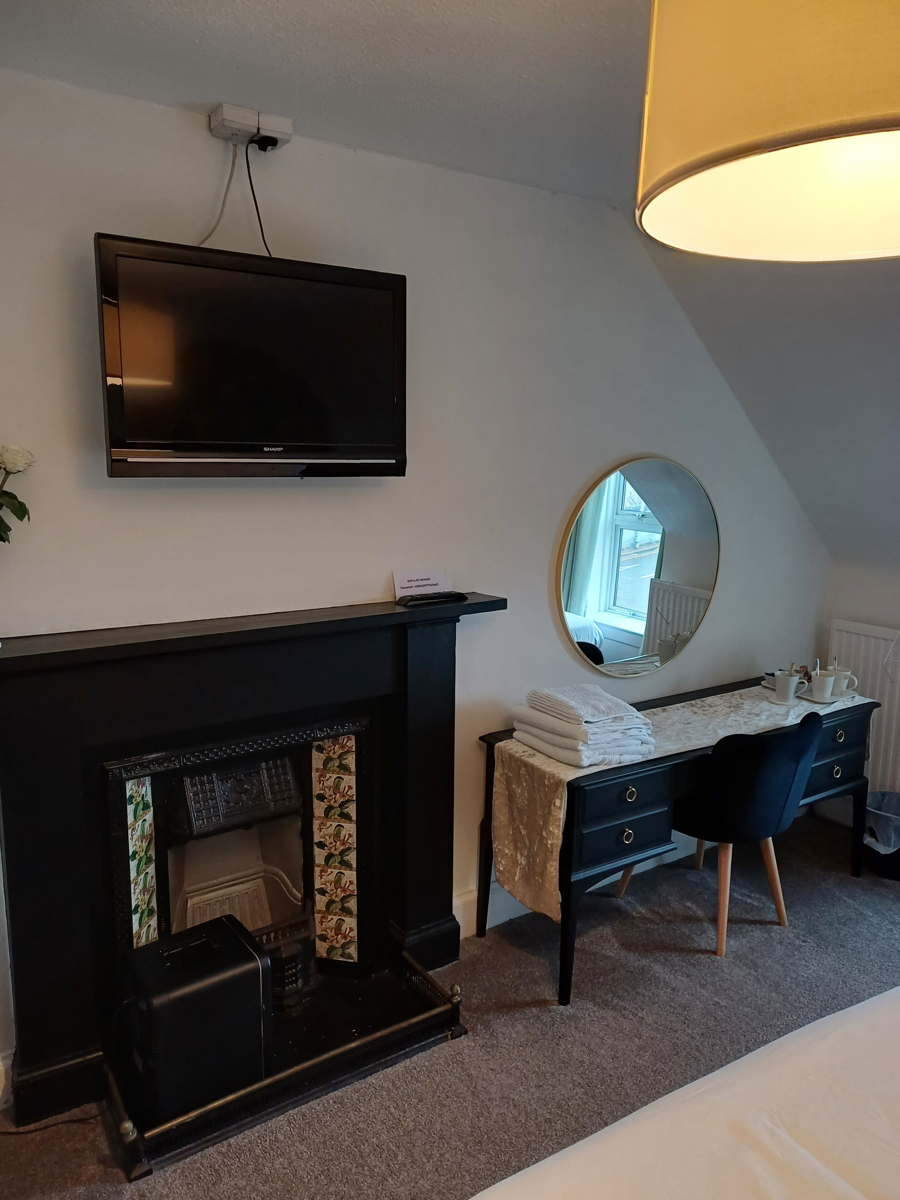 Une chambre d’hôtel moderne dotée d’un téléviseur à écran plat fixé au mur, d’une cheminée agrémentée d’un manteau décoratif, d’un bureau avec un miroir et d’une grande fenêtre laissant entrer la lumière naturelle.