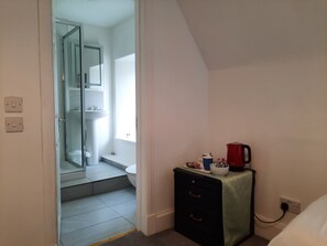 A room with a bed, a bedside table with a red kettle, a small cabinet, and a door leading to a bathroom.