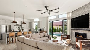 A modern kitchen with a central island, a dining area with chairs, a large beige sofa, and a fireplace. - Desert Oasis: Pool, Spa, Golf Sim & Family Fun! (Bermuda Dunes)