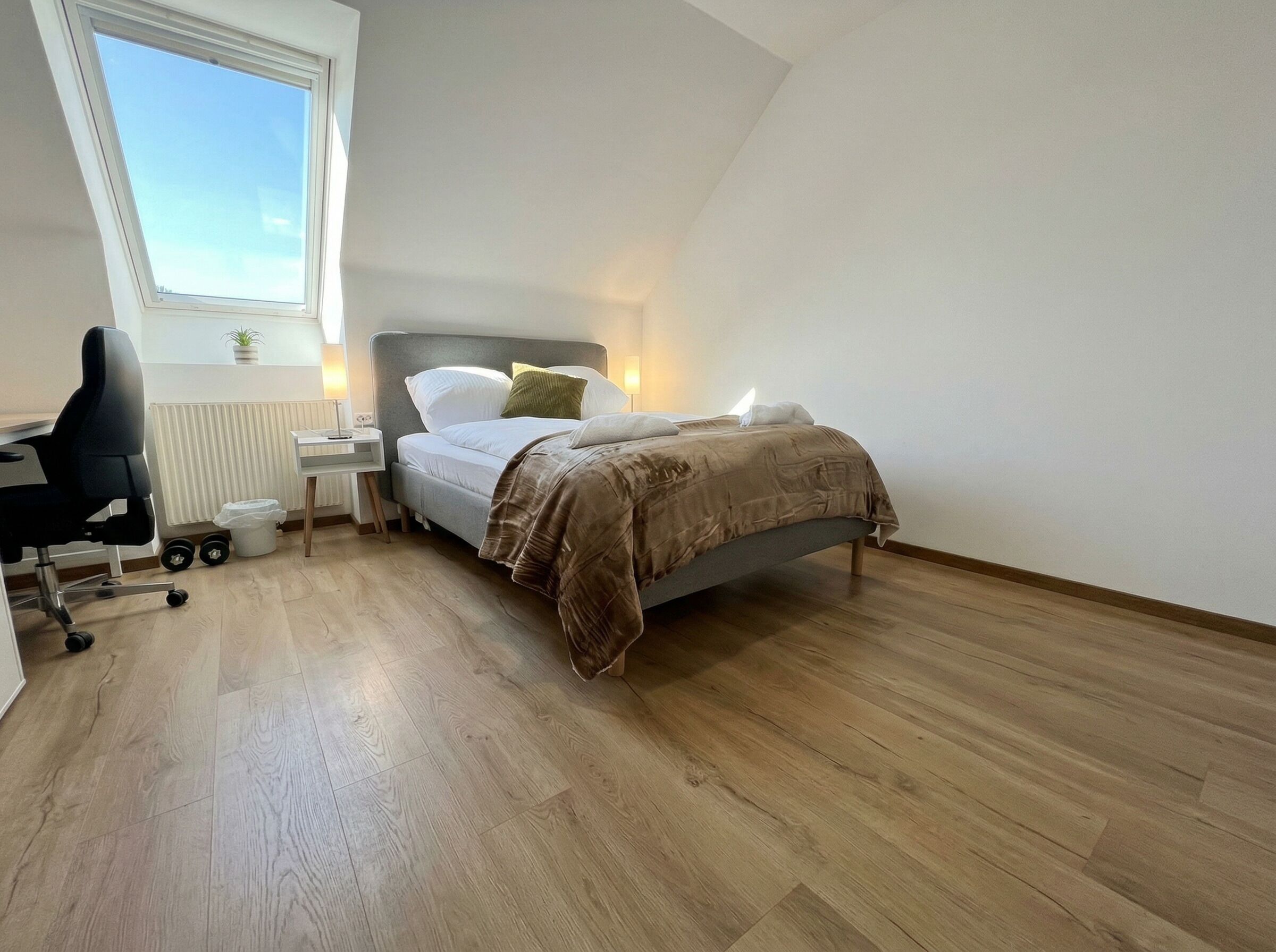 A bedroom with a bed, bedside table, lamp, chair, radiator, window, and wooden flooring.