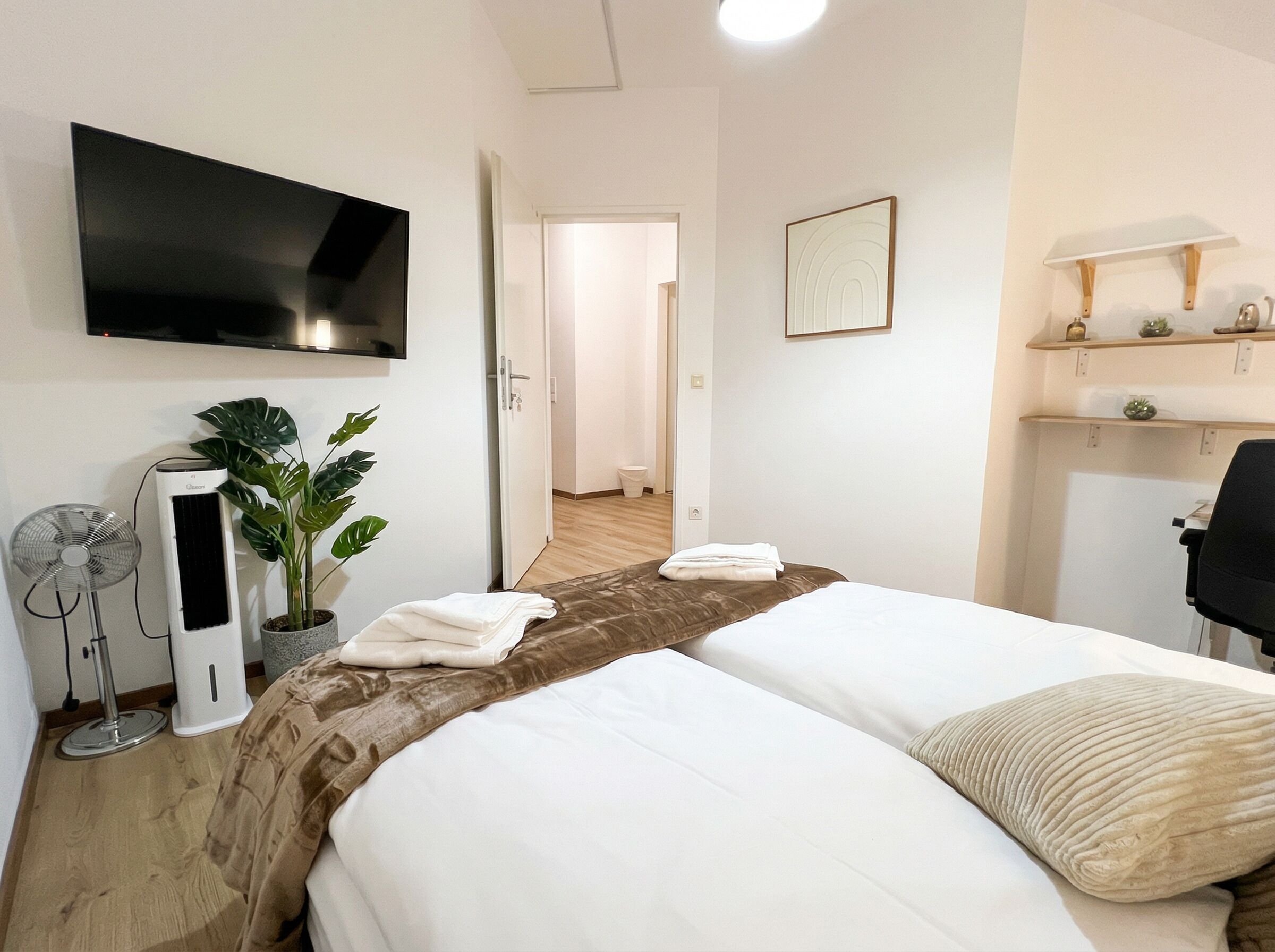 Une chambre d’hôtel avec deux lits, une télévision, une plante, un ventilateur et un bureau avec une lampe.