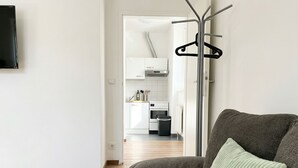 A modern living room with a grey sofa and a green pillow, a coat rack, and a view into a kitchen with white cabinets and a stove. - Lovely 3BR Apt With Balcony Rennweg (Vienna)