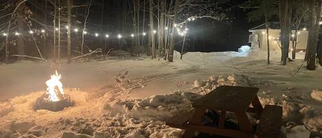 A snowy outdoor area with a campfire, a tent, and string lights.