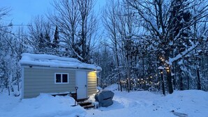 A small, single-story cabin with a snow-covered roof, surrounded by a forested area.