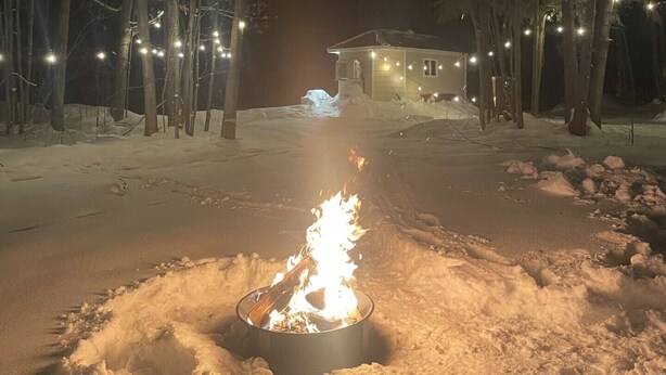 A snow-covered outdoor area with a campfire in the center, surrounded by trees and string lights.