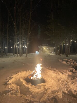 Un espace extérieur enneigé, avec un feu de camp au centre, entouré d’arbres et illuminé de guirlandes lumineuses.