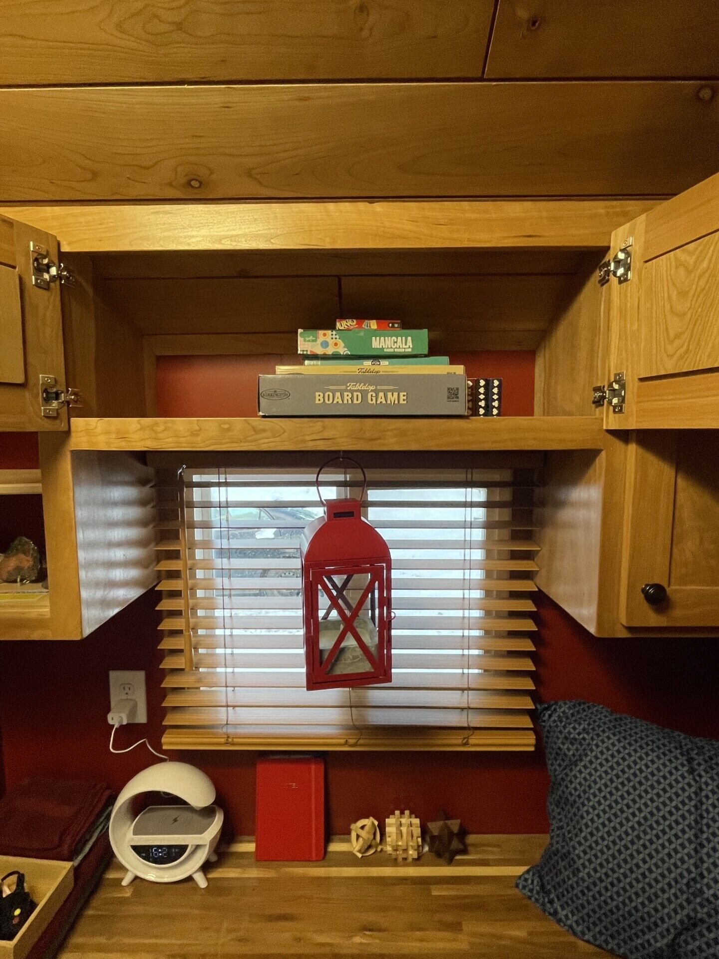 A compact room with a wooden ceiling, a window with blinds, a red lantern, and a shelf with board games.