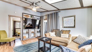 Una sala de estar con un sofá, sillones, una mesa de centro y un televisor. Hay una estantería y una ventana con cortinas.