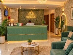 A reception area with a green counter, a laptop, and a person working. There are plants, a round table, and a green sofa. - High Five Hotels (Assagao)