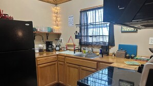 Une cuisine avec des meubles de rangement en bois, un réfrigérateur noir, une machine à café, un mixeur et une fenêtre avec un rideau.