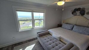 Una camera da letto con un letto grande, una panca, una ventola a soffitto e una finestra vista strada e vegetazione.