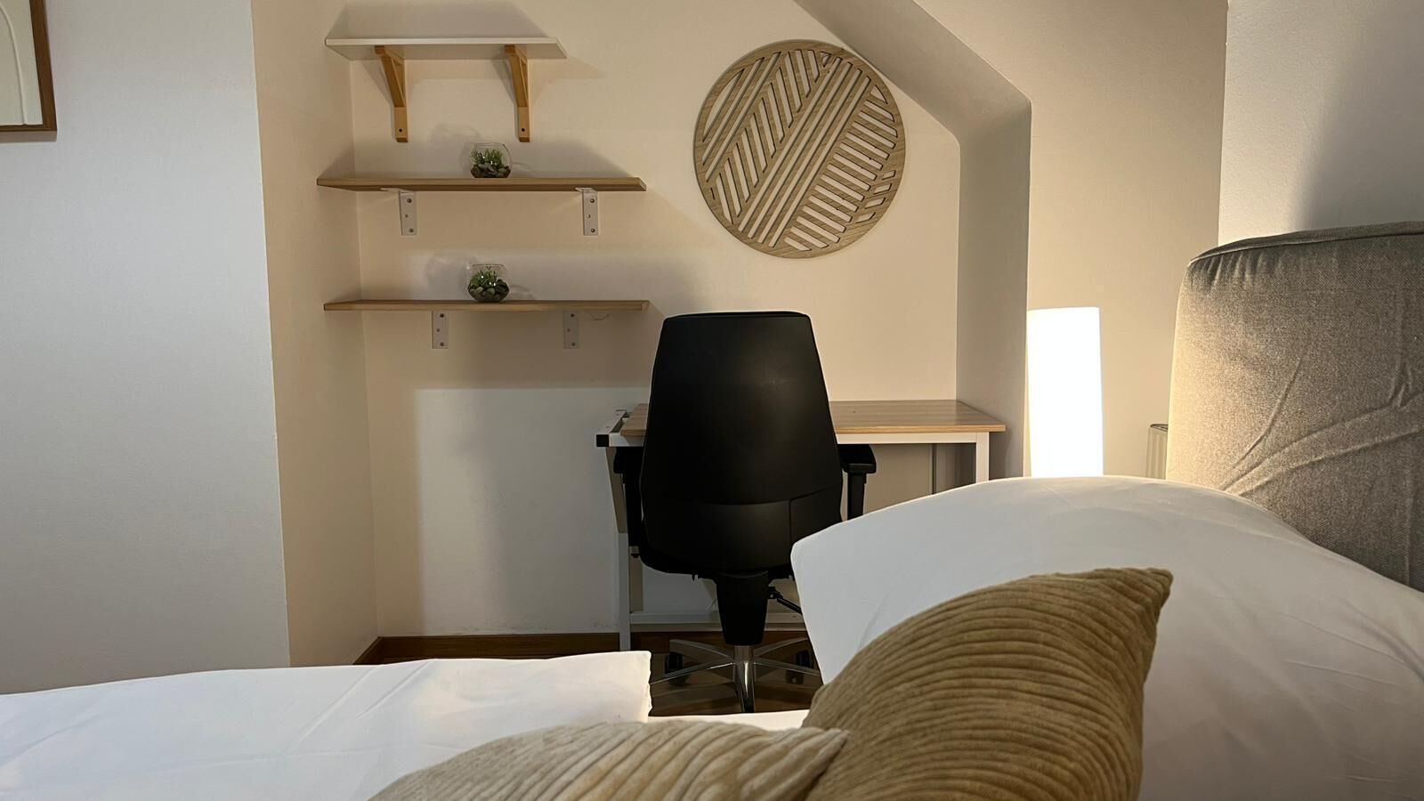 A bedroom with a bed, a desk, and a chair. There are shelves above the desk and a decorative wall piece.