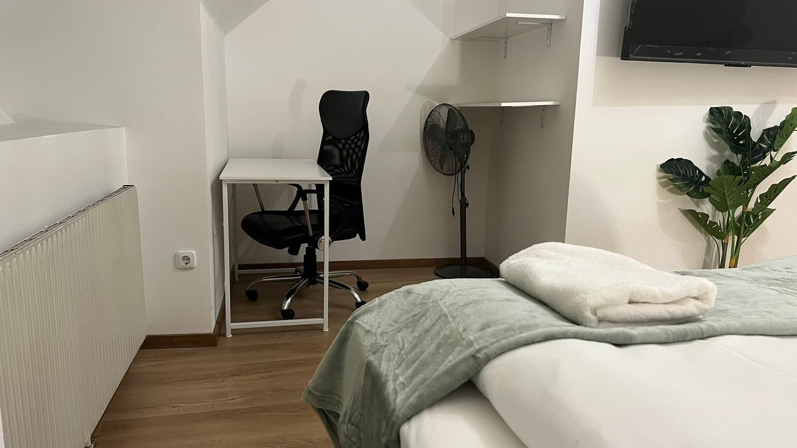 Ein Schlafzimmer mit Bett, Schreibtisch, Stuhl, Standventilator und einer Topfpflanze.