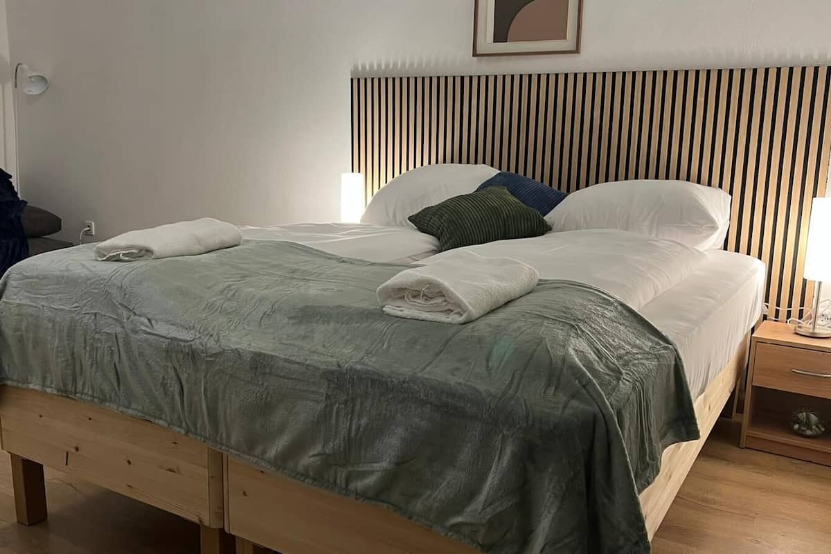 A neatly made bed with a grey blanket, a bedside lamp, and a small wooden nightstand with a drawer.