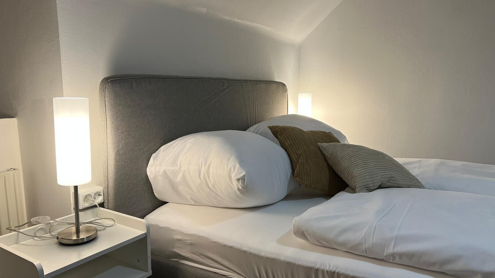 A neatly made bed with white bedding and two pillows, a bedside lamp, and a power outlet on a white nightstand.