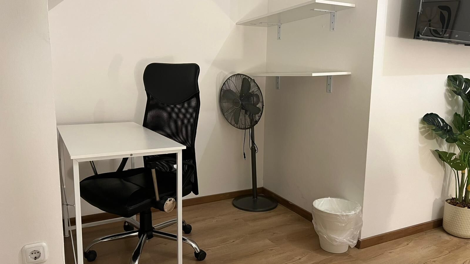 Ein kleiner Arbeitsraum mit einem weißen Schreibtisch, einem schwarzen Bürostuhl, einem Standventilator, einer weißen Mülleimer und einer Topfpflanze.