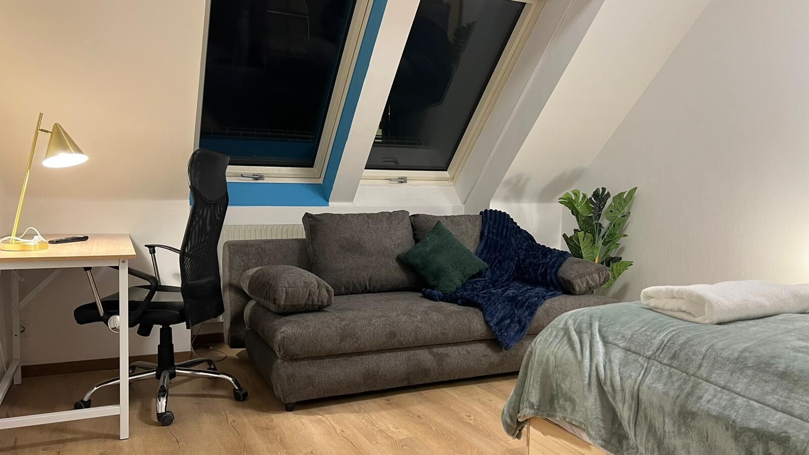 Une pièce de taille modeste, comprenant un bureau, un canapé gris, un lit et une fenêtre avec des stores.