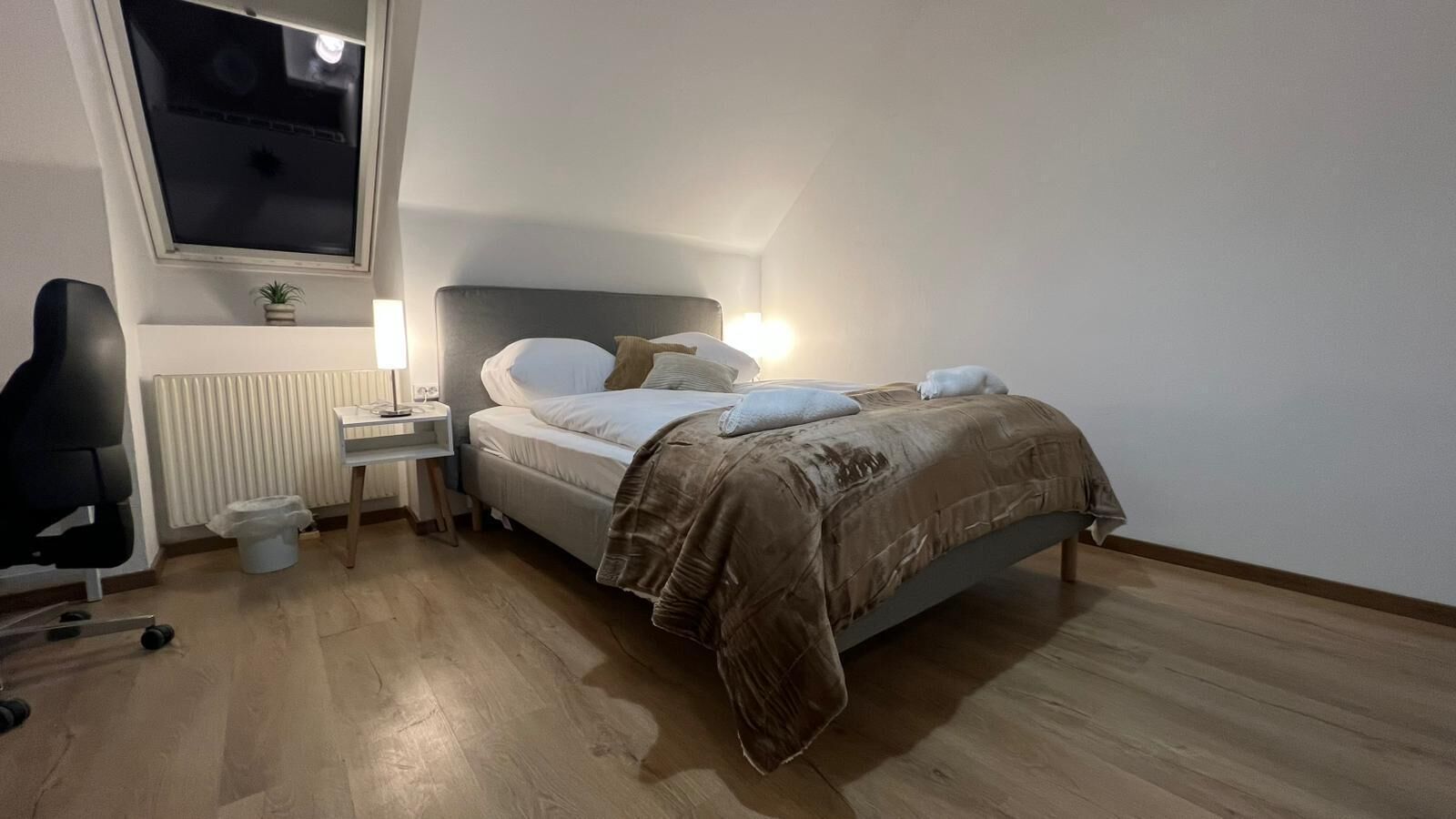 Een slaapkamer met een bed, nachtkastje, lamp, stoel, radiator en een raam.