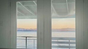 Ein Zimmer mit einem Bett und einem großen Fenster mit Blick auf ein Gewässer und ferne Berge.