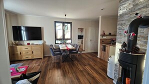 A spacious living area with a wooden floor, a dining table with chairs, a flat-screen TV, a fireplace with decorations, and a kitchen area with a window. - Small apartment on the outskirts of Berlin (Teltow)