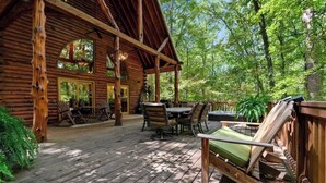 Una cabaña de madera con terraza, asientos al aire libre y un ventanal grande con vista a un bosque.