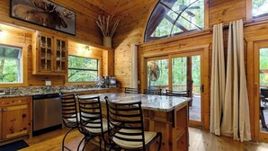 Interior de una cabaña de madera con cocina, comedor y amplios ventanales.