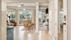A spacious living area with a large window, a ceiling fan, and a view of palm trees. - Holmes Beachfront Oasis (Holmes Beach)