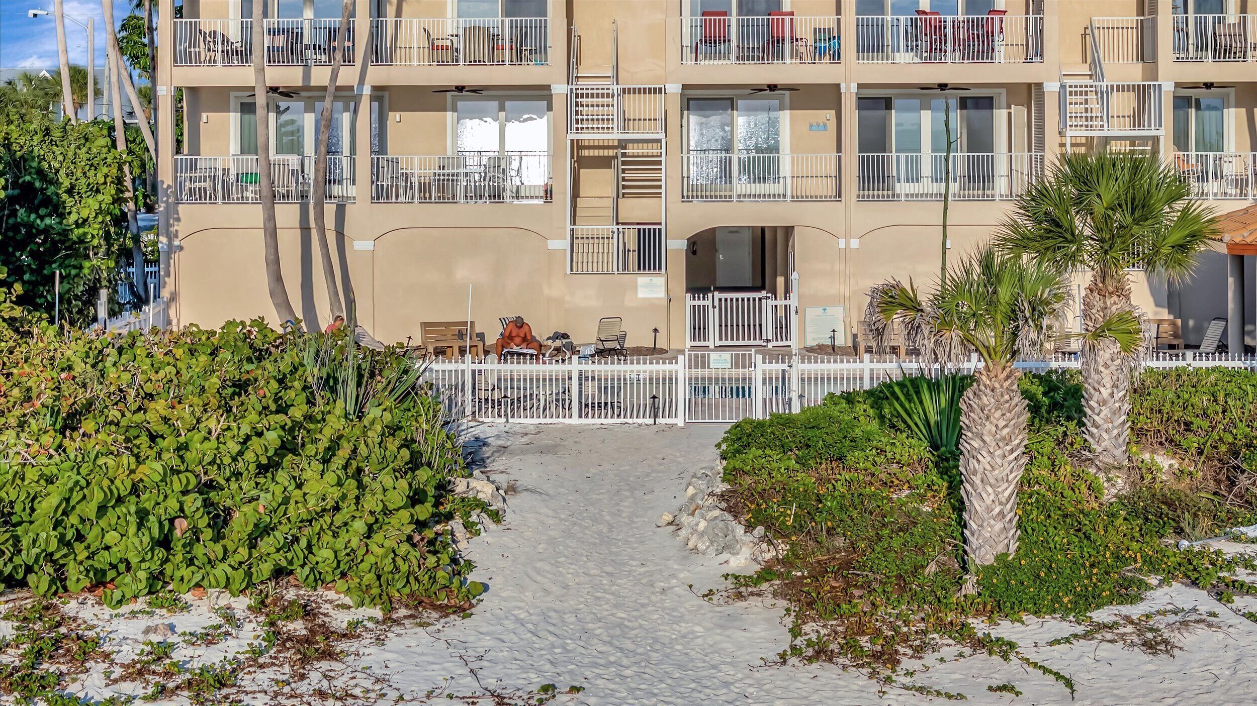 Un hotel de varios pisos con balcones, una zona de estar al aire libre techada y un camino pavimentado que lleva a la entrada.
