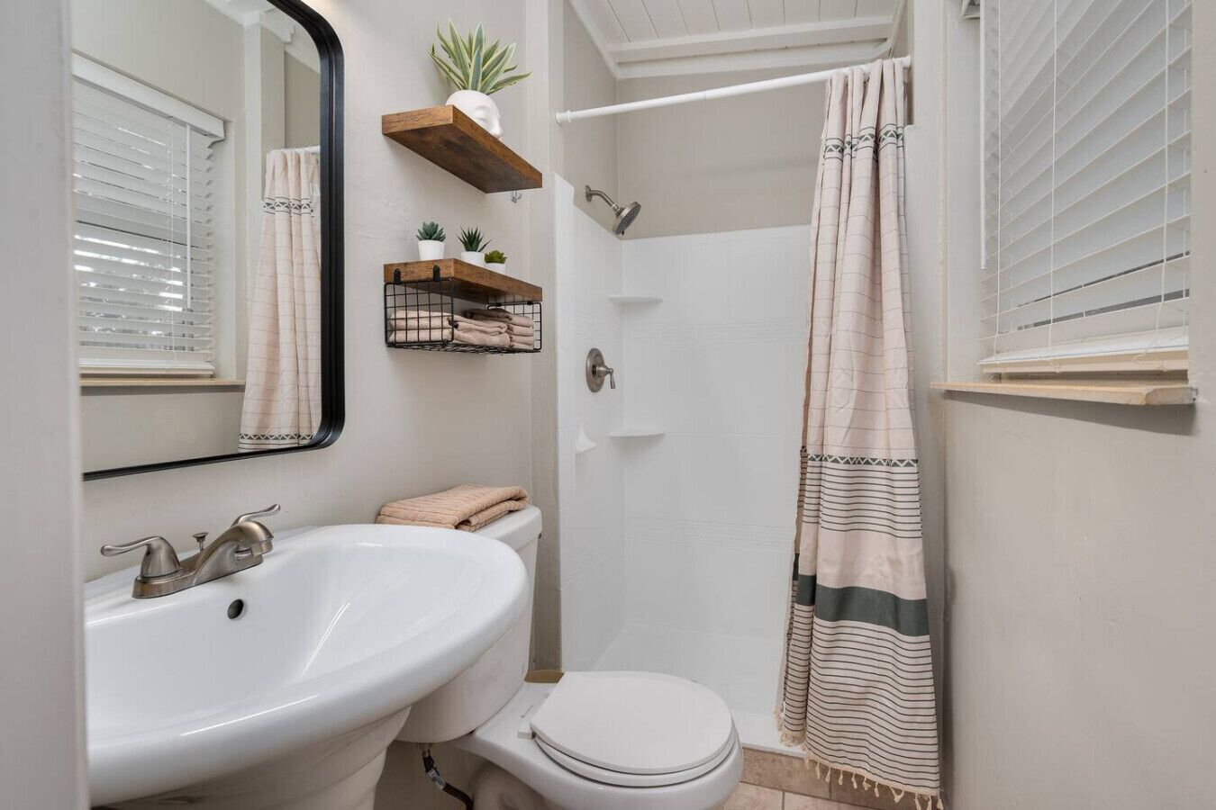 Ein Badezimmer mit weißem Waschbecken, Toilette und Dusche. Holzregale mit Pflanzen und Handtüchern sind ebenfalls vorhanden.