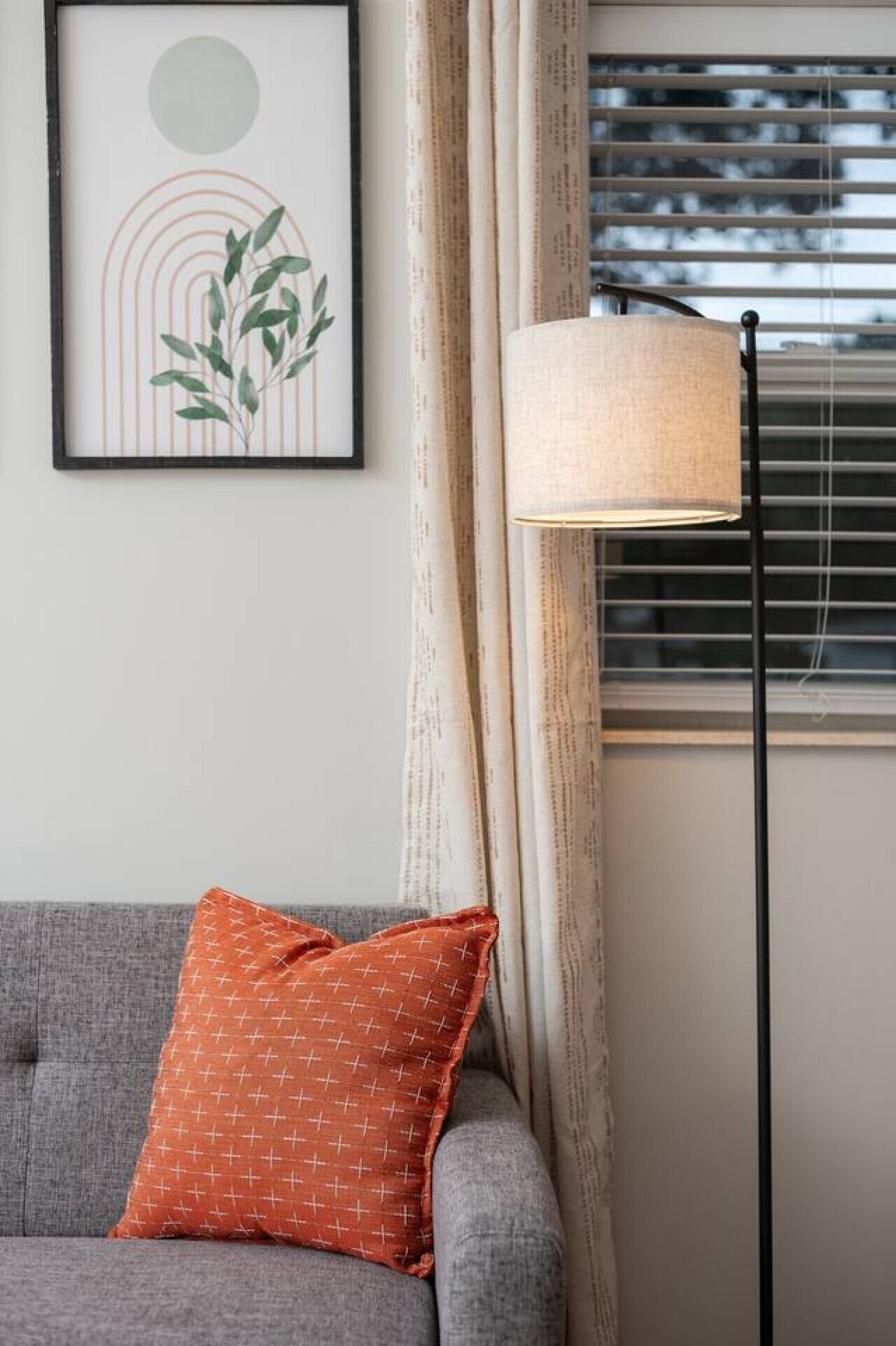 Un sofá gris con un cojín naranja estampado, una lámpara de pie y una obra de arte enmarcada en la pared.