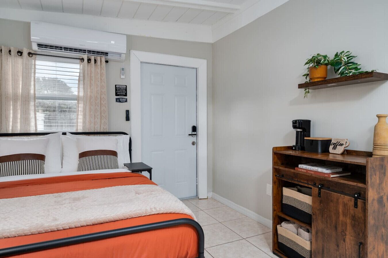 A bedroom with a bed, a desk, a chair, a shelf with books and a coffee maker, and a door.