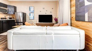 A modern living room with a white sectional sofa, a flat-screen TV, and a kitchen area with dark cabinets and stainless steel appliances. - Houston Private Retreat, 17 min Downtown (Houston)