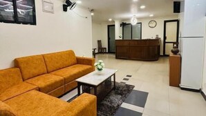 A modern living room with a sofa, a coffee table, and a rug. There is a reception desk in the background. - HOTEL IHAM PARADISE (Mangaluru)