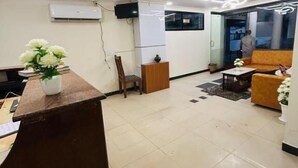 A modern reception area with a wooden reception desk, a seating area with a sofa and armchairs, and a television mounted on the wall. - HOTEL IHAM PARADISE (Mangaluru)