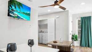 A modern kitchen with white appliances, a dining area with a wooden table and two wine glasses, and a TV displaying a tropical beach scene. - Cozy 1-bedroom apartment in beautiful Gulf Breeze with AC (Gulf Breeze)