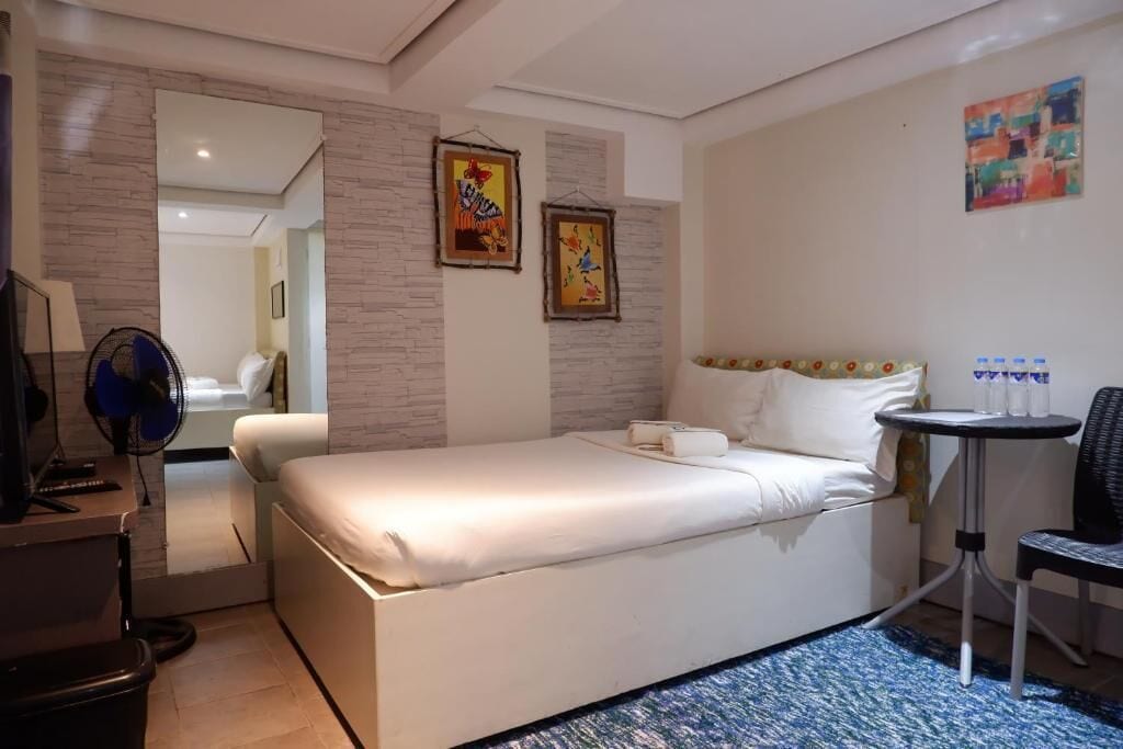 A hotel room with a bed, a chair, a small table with water bottles, and two wall-mounted paintings.