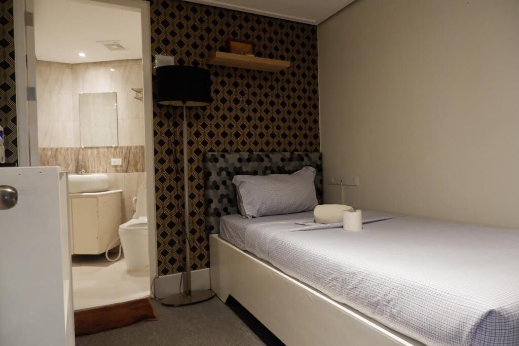 A small, compact room with a single bed, a bedside table, a floor lamp, a toilet, and a mirror.