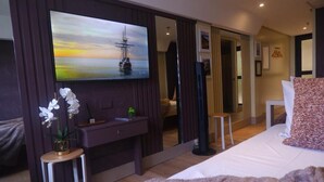 A hotel room with a bed, a large mirror, a TV on the wall, a small table with a plant, and a nightstand.