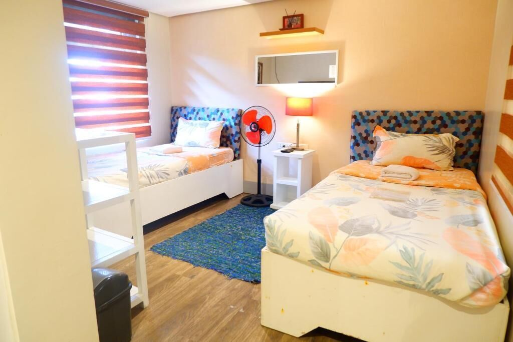 Un dormitorio con dos camas, un ventilador de pie, una mesita de noche, un espejo y una ventana con persianas.