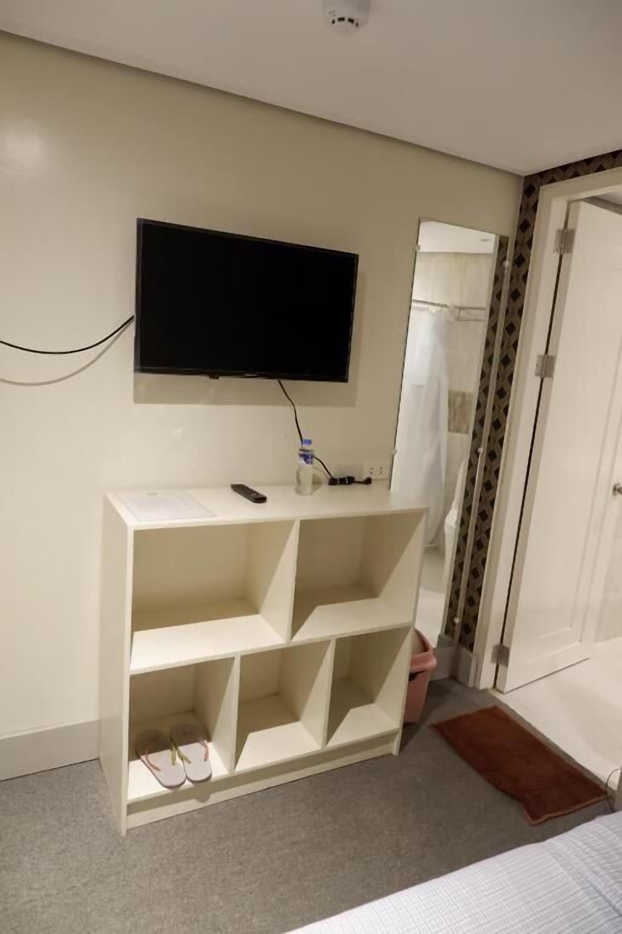 A room with a TV mounted on the wall, a white console table with shoes, a door, and a shower area.