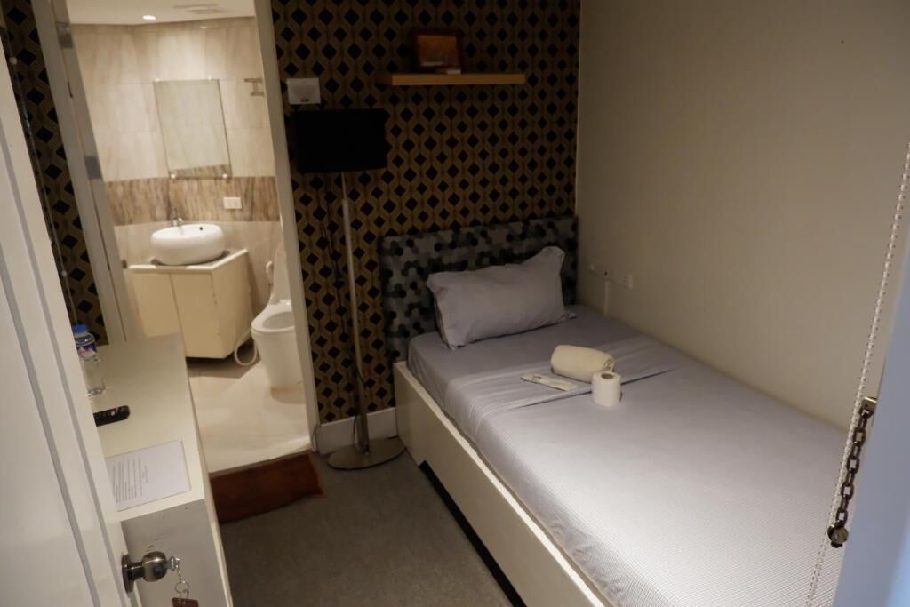 A small, compact room with a single bed, a bedside table, a nightstand, a wall-mounted shelf, a bedside lamp, and a bathroom with a sink and mirror.
