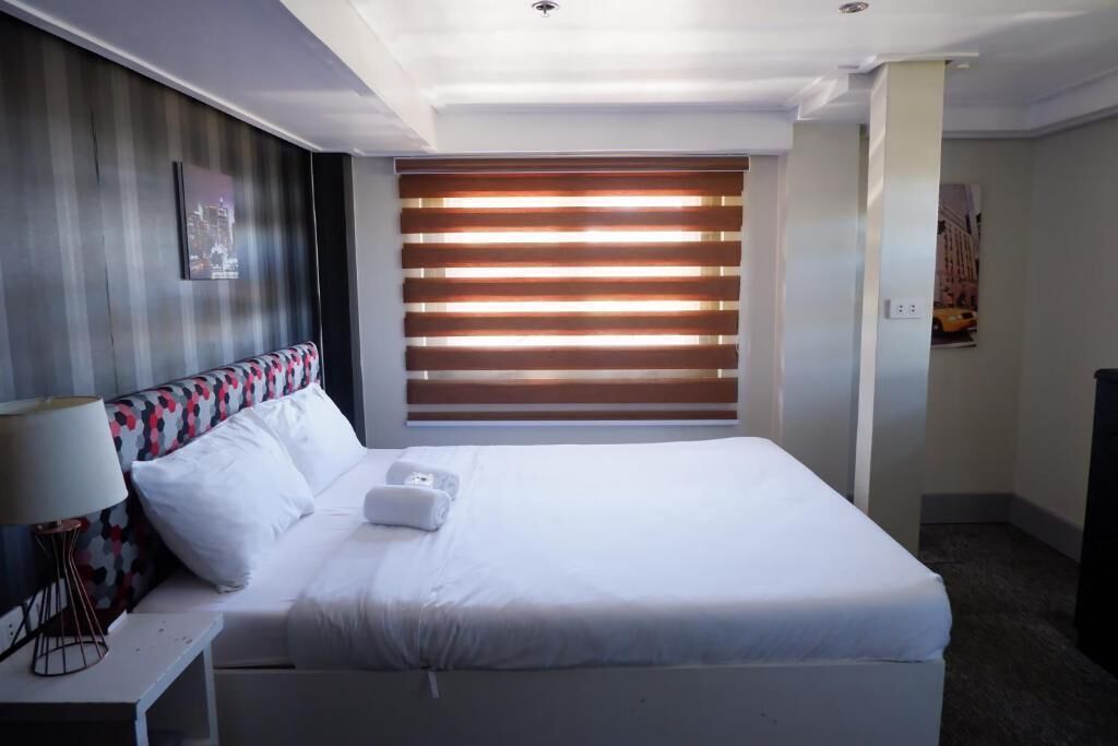 A hotel room with a bed, a bedside table with a lamp, a wall-mounted TV, and a window with blinds.