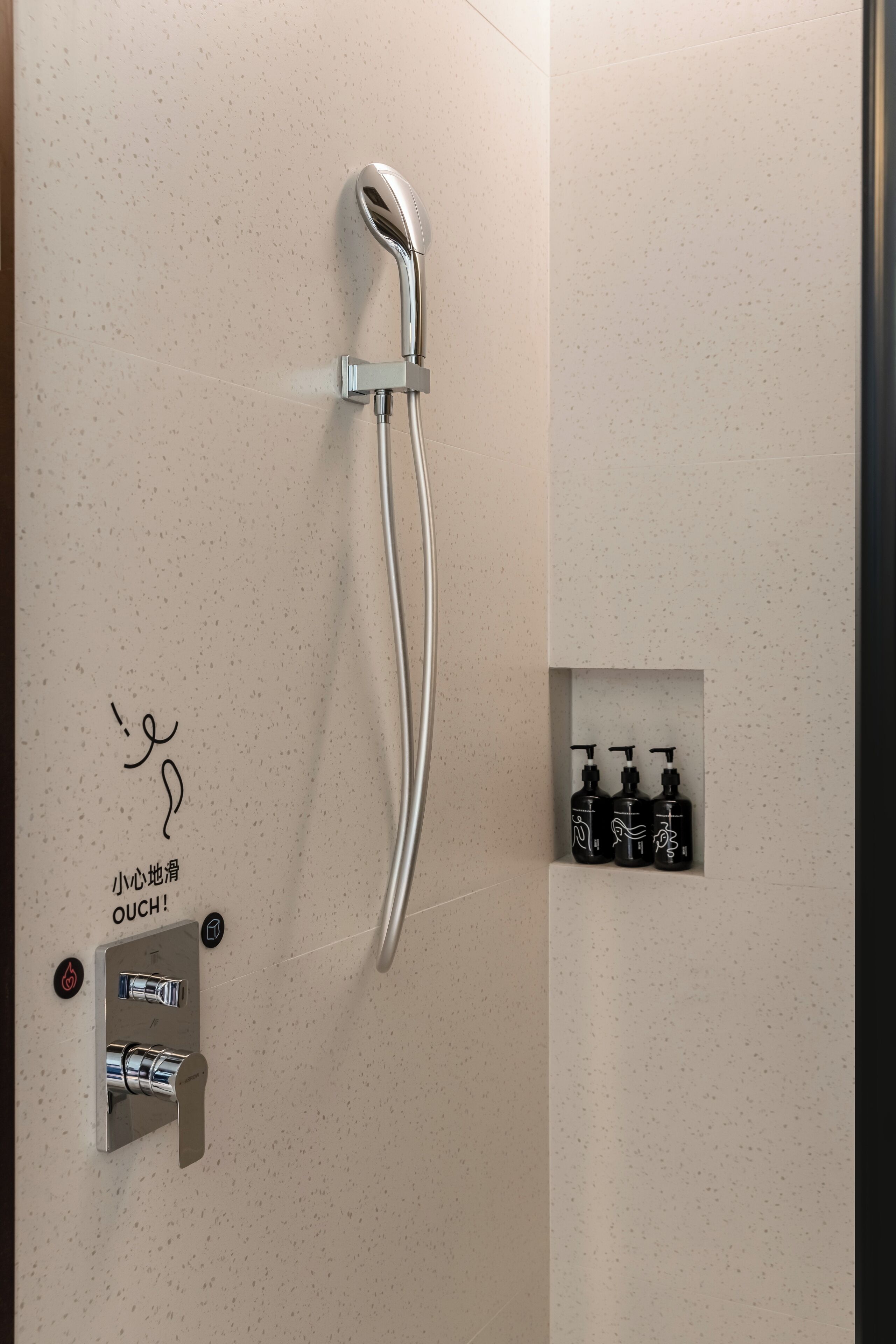 A shower with a handheld design, a control panel with a temperature knob, and three black bottles of shampoo and conditioner on the wall.
