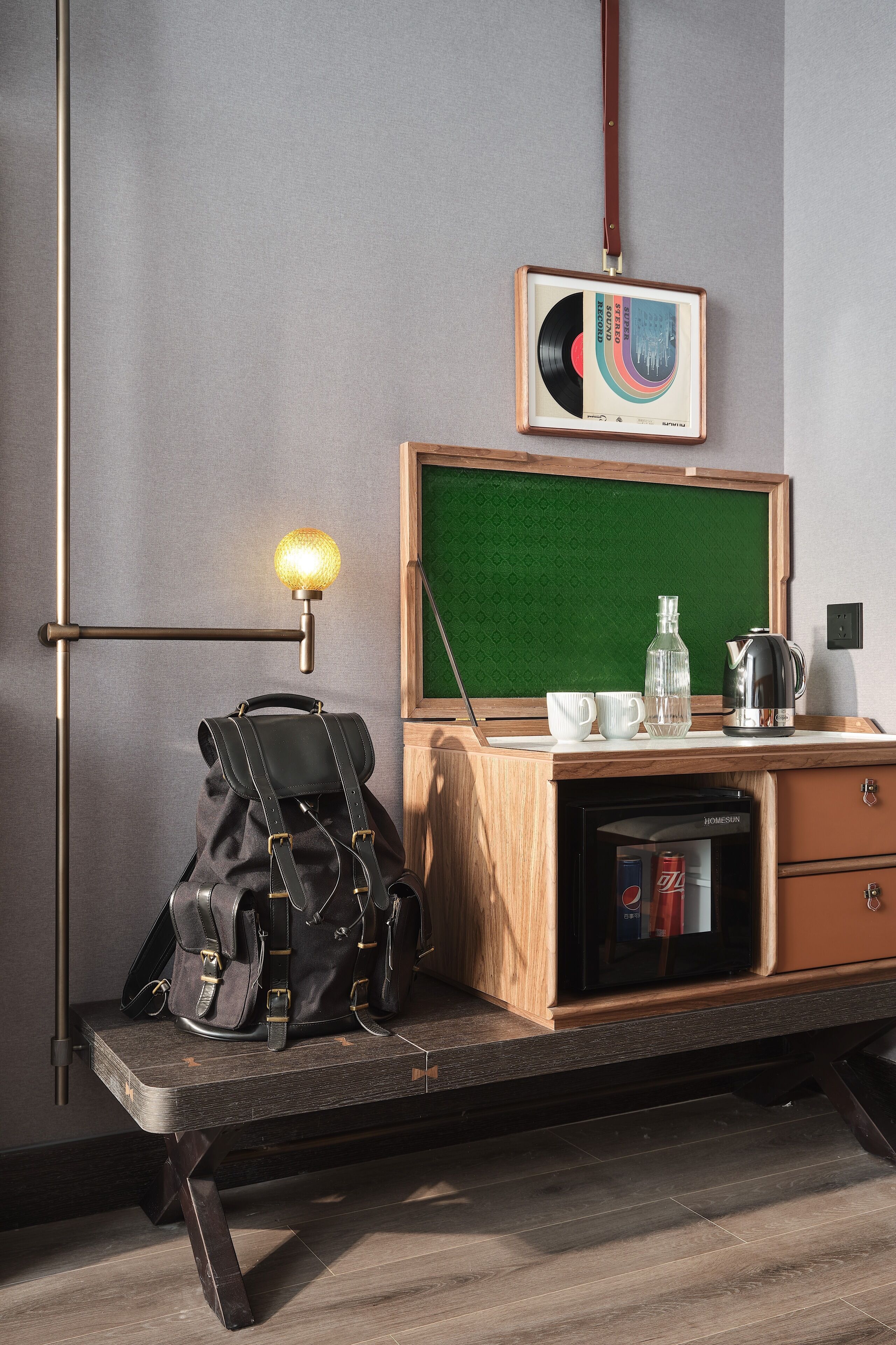 A room with a desk, a green chalkboard, a framed vinyl record, a lamp, a backpack, a water bottle, a coffee pot, and a refrigerator.